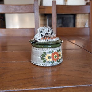 Pode incluir: Um pote de cerâmica com tampa, com um design floral em laranja, amarelo e verde. O pote tem uma base cinza claro com uma borda decorativa e uma borda verde. A tampa tem uma alça decorativa.