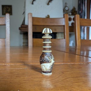 Könnte beinhalten: Kleine, verzierte, braun-cremefarbene Dekoflasche. Sie zeigt eine detaillierte Hirschgravur und ein gestapeltes, zylindrisches Design. Der Deckel hat die Form eines Pilzes. Die Flasche ist etwa 10 cm hoch.