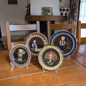May include: Four decorative ceramic plates with folk art designs. The plates feature brown, blue, and yellow rims, and depict figures and floral patterns. The plates are displayed on clear stands on a wooden table.