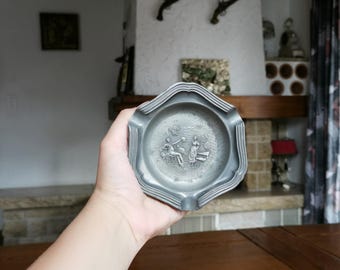 May include: A pewter ashtray with a scalloped edge, held in a hand. The ashtray features a detailed relief scene of figures in a garden. The ashtray is a muted gray color and appears to be vintage.