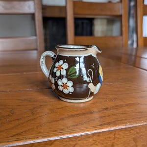 May include: A small, brown ceramic creamer with a white handle and spout. The creamer is decorated with white flowers with orange centers and green leaves. A cartoon figure is painted on the side. The creamer sits on a wooden table.