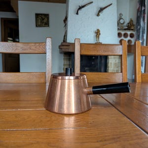 Puede incluir: Una pequeña cacerola de cobre con un asa negra y un asa de tapa de madera está sobre una mesa de madera. La cacerola tiene forma cónica y un pico. El fondo incluye una chimenea y sillas de madera.