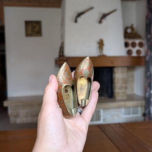 May include: A pair of small, decorative brass shoes with intricate floral and leaf patterns in red, orange, and green. The shoes are held in a hand, showcasing their detailed craftsmanship and vintage aesthetic. The interior of the shoes is visible.