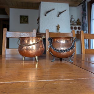 Op de afbeelding: Twee koperen ketels met gehamerde texturen staan op een houten tafel. Eén heeft messing poten en een touwgreep, de andere heeft een kettinggreep en smeedijzeren poten. De achtergrond omvat houten stoelen en een open haard.