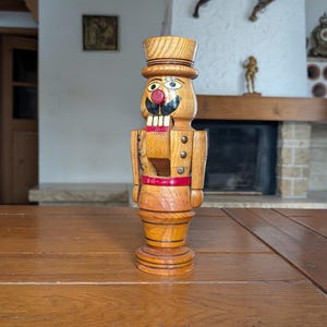Peut inclure: Figurine de casse-noix en bois avec un chapeau haut de forme, un nez rouge et une moustache noire. La figurine a un design détaillé avec une ceinture rouge et des boutons dorés. Le casse-noix est sur une table en bois.