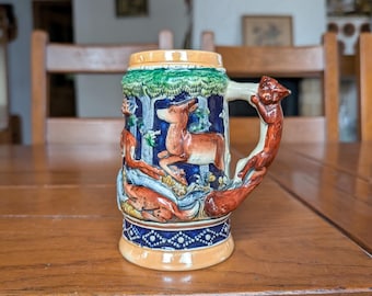 Puede incluir: Un jarro de cerveza de cerámica con una escena de bosque con un ciervo y zorros. El asa tiene forma de zorro. El jarro tiene una combinación de colores azul, verde y marrón con patrones decorativos alrededor de la base y el borde.