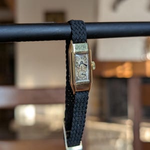 May include: A vintage gold-toned rectangular wristwatch with a black woven band. The watch face has a light-colored background with black numerals and hands. The watch is suspended from a black metal bar.