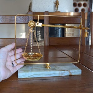 Könnte beinhalten: Eine dekorative goldfarbene Waage mit rechteckigem Rahmen und einer kleinen Schale, die an Ketten hängt. Die Waage steht auf einem rechteckigen, marmorähnlichen Sockel. Die Waage ist ein dekorativer Gegenstand.
