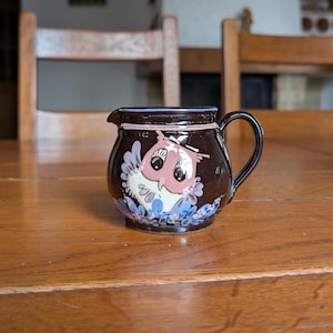 May include: A small, brown ceramic creamer with a black handle and a spout. The creamer features a hand-painted owl design in pink, white, and blue. The rim is trimmed with a blue and pink floral pattern. A charming addition to any kitchen.