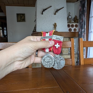 Puede incluir: Dos medallas de plata con cintas rojas y blancas, una con una cruz suiza. Las medallas presentan grabados detallados, uno con un grupo de figuras y el otro con una sola figura. Las cintas están unidas a una barra con el texto "1964".
