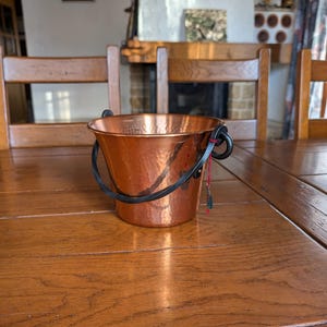 Op de afbeelding: Een kleine, gehamerde koperen emmer met een zwarte handgreep en een rood-groen decoratief accent. De emmer staat op een houten tafel. De emmer is een decoratief item.