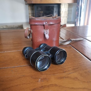 May include: Black binoculars with gold-colored trim rest on a wooden table next to a brown leather case with a shoulder strap. The case has a buckle closure and a handle. The background includes a fireplace and a window.