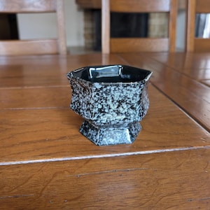 May include: A black and white speckled ceramic bowl with a hexagonal shape. The bowl has a pedestal base and is sitting on a wooden table. The speckled pattern is a mix of black and light blue.