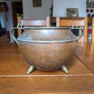 May include: A copper cauldron with brass handles and feet. The pot has a rounded body and a wide rim. The copper has a warm, aged patina. The cauldron is sitting on a wooden table.