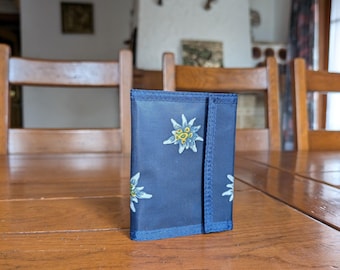 May include: A navy blue wallet with a flap closure, featuring a repeating pattern of white and yellow edelweiss flowers. The wallet is sitting on a wooden table.