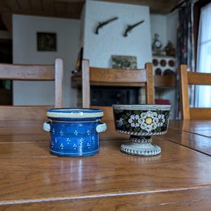 Pode incluir: Dois objetos decorativos de cerâmica sobre uma mesa de madeira. À esquerda, um pote azul com pontos brancos e pequenas alças. À direita, uma xícara com design floral em preto e branco com base.