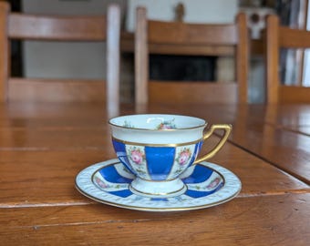 Może przedstawiać: Zestaw filiżanki i spodka do herbaty w stylu vintage. Filiżanka i spodek mają niebiesko-biały wzór z kwiatowymi akcentami i złotymi obramowaniami. Filiżanka ma złoty uchwyt. Zestaw jest umieszczony na drewnianym stole.
