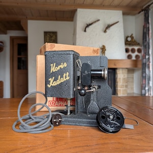 Puede incluir: Un proyector de películas Noris Kadett negro vintage con un cable de alimentación gris enrollado y una bobina de película. El proyector tiene el texto "Noris Kadett" en la parte delantera. El proyector está sobre una mesa de madera.