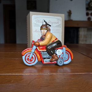 May include: A vintage toy motorcycle with a rider. The motorcycle is red with blue and yellow accents. The rider wears a brown helmet and jacket. The number 15 is on the side of the motorcycle. A box with text is in the background.