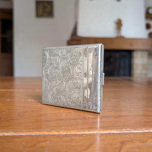 Könnte beinhalten: Eine silberne Zigarettenetui mit einem verzierten floralen Muster. Das rechteckige Etui hat einen Klappdeckel und einen Verschluss. Es liegt auf einer Holzoberfläche. Das Etui ist wahrscheinlich Vintage.