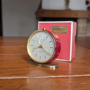 May include: A vintage Looping Swiss alarm clock with a gold-tone face and hands, set on a gold base. The clock has a red and gold casing and is accompanied by its original red box. The clock face displays the Looping Swiss logo.