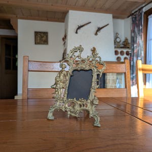 May include: Ornate antique brass picture frame with a black insert. The frame features detailed scrollwork, a small figure on the side, and a decorative top. It is displayed on a wooden table.