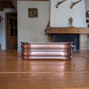 Puede incluir: Una jardinera rectangular de cobre con un diseño acanalado y asas de metal azul. La jardinera está sobre una mesa de madera. El cobre tiene un color marrón rojizo cálido.