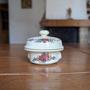 以下が含まれることがあります： 赤、青、緑の花柄が施された、クリーム色の蓋付きセラミック皿。木製の表面に置かれています。蓋の上部には小さなノブが付いています。花柄は皿の底にも繰り返されています。