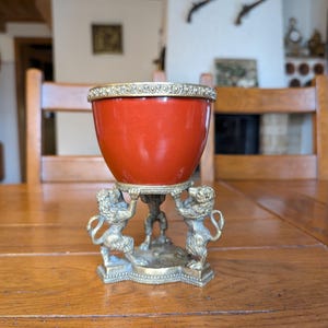 Puede incluir: Un recipiente rojo en forma de huevo con un borde dorado decorativo, sostenido por una base de latón con tres figuras de león. El recipiente se encuentra sobre una mesa de madera, mostrando una estética vintage.