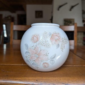 Puede incluir: Un jarrón de cerámica blanca con forma redonda, adornado con un estampado floral en tonos melocotón, rosa y gris. El jarrón está sobre una mesa de madera. El diseño floral incluye flores grandes y pequeñas y hojas.