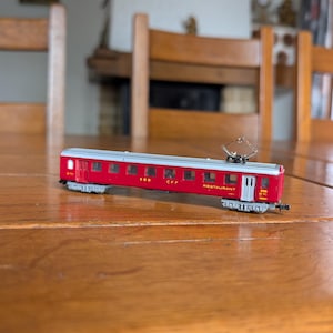 May include: A red and silver model train car labeled "SBB CFF RESTAURANT" on a wooden table. The miniature train car has detailed windows and a pantograph on top. The background includes wooden chairs and a fireplace.