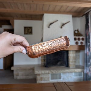 Puede incluir: Una sartén rectangular de cobre con textura martillada. La sartén tiene un borde oscuro y asas pequeñas en cada extremo. El cobre tiene un color marrón rojizo cálido. La sartén se sostiene frente a una chimenea.