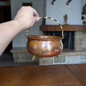 May include: A small, copper-colored cauldron with a brass handle and three small feet. The cauldron has a rounded body and a shiny, reflective surface. The handle is arched and twisted. The cauldron is held up by a hand.