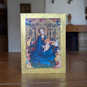 以下が含まれることがあります： 聖母子像を描いた額入りの宗教芸術作品。 金色のフレームで、青いガウンを着て幼子イエスを抱く聖母マリアが描かれています。 天使や他の人物が彼らを囲み、詳細な背景があります。