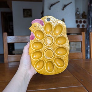 Puede incluir: Un portahuevos de cerámica amarilla con forma de gallina, con una cresta rosa y múltiples compartimentos ovalados para huevos. El soporte tiene una superficie texturizada y un agujero para colgar. Un artículo de cocina decorativo.