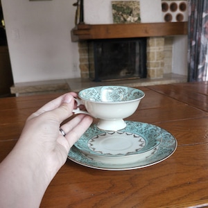 May include: A vintage tea set comprising a teacup, saucer, and side plate. The teal and gold floral pattern adorns the white-based teacup and plates. The set is presented on a wooden table, highlighting the elegant design.