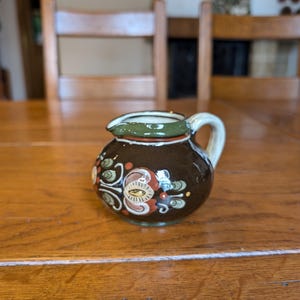 Pode incluir: Um pequeno cremeira de cerâmica marrom e redonda com uma alça branca e uma borda verde. A cremeira é decorada com padrões florais coloridos em vermelho, laranja e branco. A cremeira está em uma mesa de madeira.