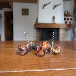 Pode incluir: Dois saca-rolhas de madeira com formas naturais repousam sobre uma mesa de madeira. Os saca-rolhas têm um esquema de cores castanho escuro e castanho claro, com um mecanismo de saca-rolhas de metal. O fundo mostra uma lareira e um interior rústico.