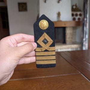 Könnte beinhalten: Eine schwarze Schulterklappe eines Marineoffiziers mit goldenen Akzenten. Sie zeigt ein goldenes Ankeremblem, ein rautenförmiges Abzeichen und goldene Streifen. Der Hintergrund ist ein Holztisch.