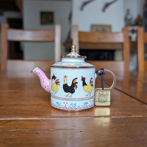 May include: A white ceramic teapot with a pink spout and handle, decorated with rooster illustrations. The teapot has a gold-coloured lid and trim, and a small tag is attached to the handle.