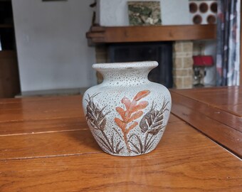 May include: A small, speckled ceramic vase with a wide base and narrow neck. The vase features a design of brown and orange leaves and foliage. The vase is sitting on a wooden table.