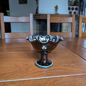 May include: A brown glass pedestal bowl with a scalloped rim. The bowl features white and green floral designs and a turquoise trim. The bowl is sitting on a wooden table.