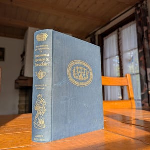 May include: A blue hardcover book, "The Concise Encyclopedia of Continental Pottery & Porcelain" by Reginald G. Haggar. The cover features gold lettering and illustrations. The book is displayed on a wooden table.