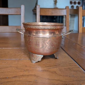 May include: A copper-coloured bowl with two handles and three decorative feet. The bowl features etched patterns around the rim and body. The bowl is sitting on a wooden table.
