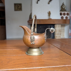 Puede incluir: Un antiguo cubo de carbón de cobre y latón con asa. El cubo tiene una cara decorativa en la parte delantera y se asienta sobre una base de latón. El asa también es de latón. El cubo está sobre una mesa de madera.