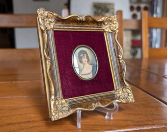 May include: Ornate gold-colored framed portrait of a woman with a burgundy velvet backing. The portrait is oval-shaped and features a woman with dark hair and a white dress. The frame is supported by a clear plastic stand, set on a wooden table.