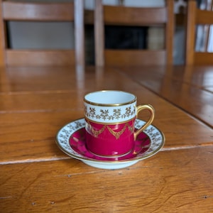May include: A small, ornate espresso cup and saucer set. The cup and saucer are a deep magenta with gold filigree and a white rim. The cup has a gold handle. The saucer is round and matches the cup's design.