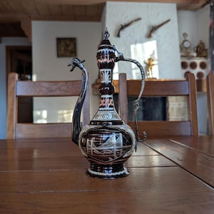 Puede incluir: Una jarra decorativa marrón oscuro adornada, con un asa y un pico plateados. La jarra presenta diseños intrincados y una ilustración de puente. Se exhibe sobre una mesa de madera, mostrando su detallada artesanía y estética vintage.