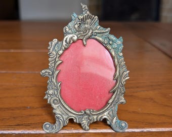 Puede incluir: Marco de fotos ornamentado de estilo antiguo, con acabado bronce y respaldo de tela roja. El marco tiene una abertura ovalada y adornos decorativos. El marco se apoya en dos pequeños pies.