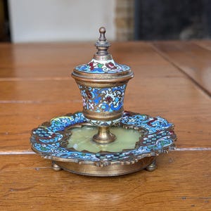 May include: An antique, ornate brass and enamel inkwell. The inkwell features a detailed floral design in blue, red, and white on a brass base. The inkwell sits on a matching tray with a scalloped edge and small feet.